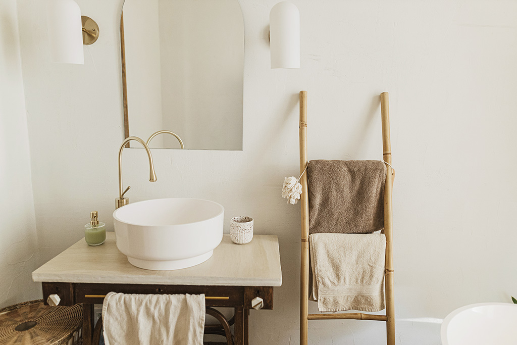 Stylish boho bathroom design. Modern ceramic sink with golden faucet on rustic stand with soap and ladder with towel on background of wall with mirror, modern bathroom interior.