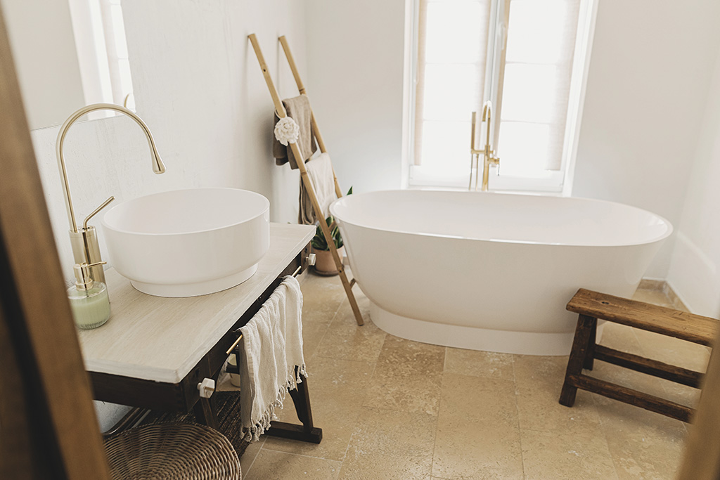 Stylish boho bathroom design. Modern bathtub with golden faucet from floor, wooden ladder with towels, ceramic sink on rustic stand and bench, modern eco bathroom interior.